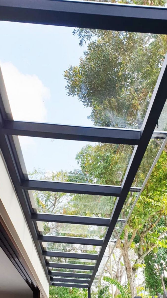 Modern black metal pergola or patio roof frame with a clear polycarbonate  canopy, showing green trees and blue sky above.