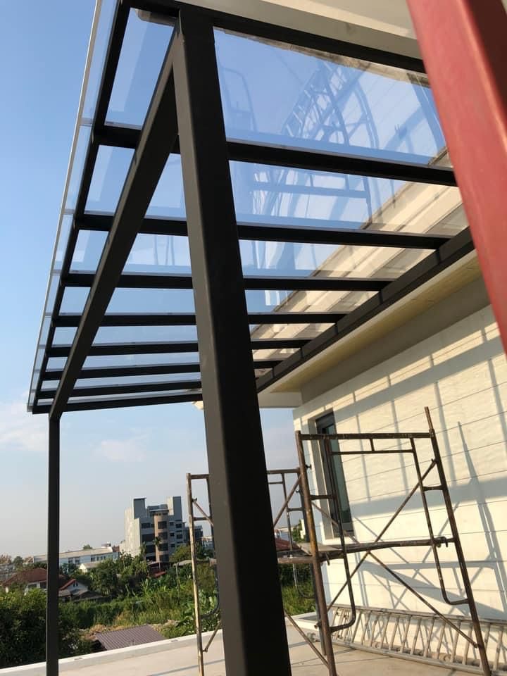 A modern black metal frame canopy with clear transparent polycarbonate sheet installed on a rooftop terrace next to a white building, with scaffolding visible underneath.