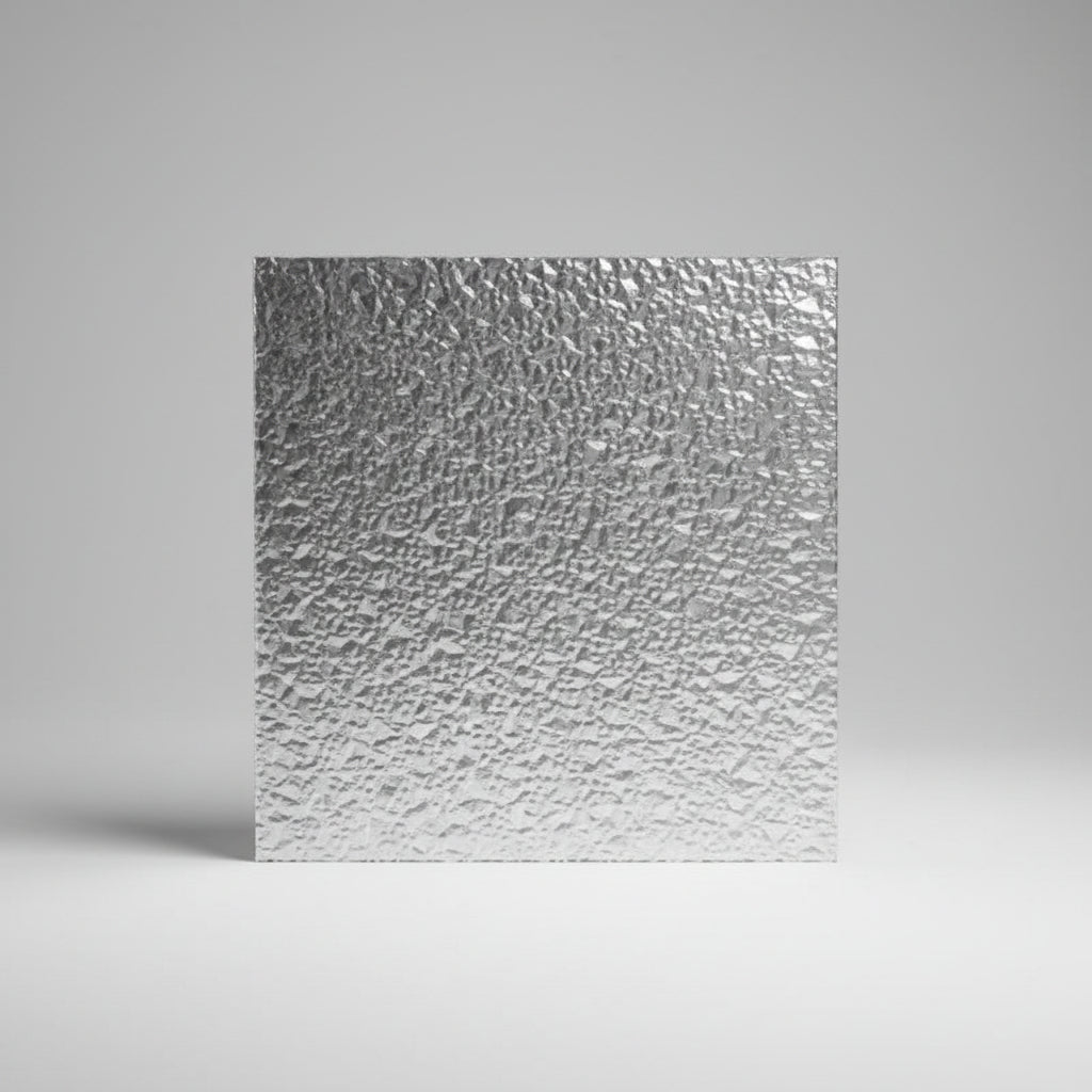 A square sheet of textured silver adhesive film or transfer paper held up in front of a blurred indoor background with windows and blinds.